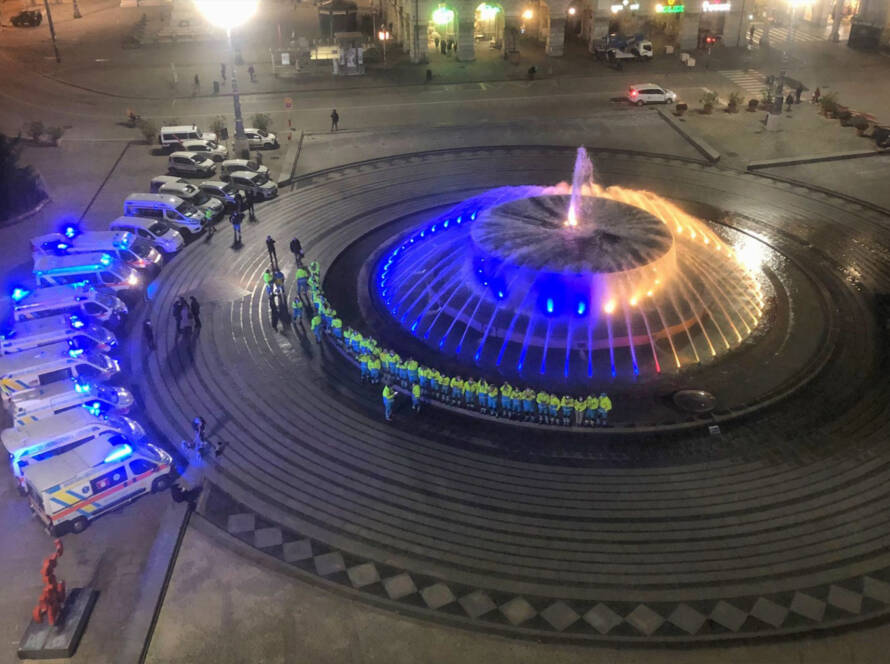 Vista dall'alto della fontana monumentale di Piazza De Ferrari a Genova, illuminata di blu e giallo in orario notturno, circondata da personale di soccorso e ambulanze disposte a semicerchio